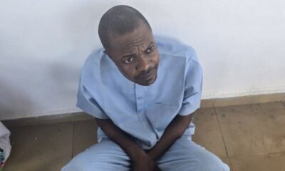 Man in light blue clothing sits on a wooden floor, looking up with a puzzled expression against a plain white wall.