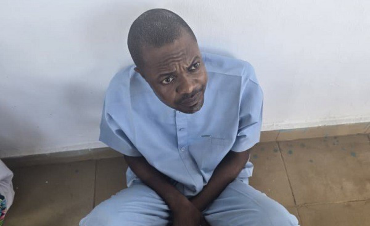 Man in light blue clothing sits on a wooden floor, looking up with a puzzled expression against a plain white wall.