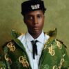 Portrait of a person wearing a green monogram jacket, white shirt, black tie, and cross necklace, with a jeweled hat motif against a green backdrop.