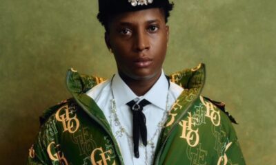 Portrait of a person wearing a green monogram jacket, white shirt, black tie, and cross necklace, with a jeweled hat motif against a green backdrop.