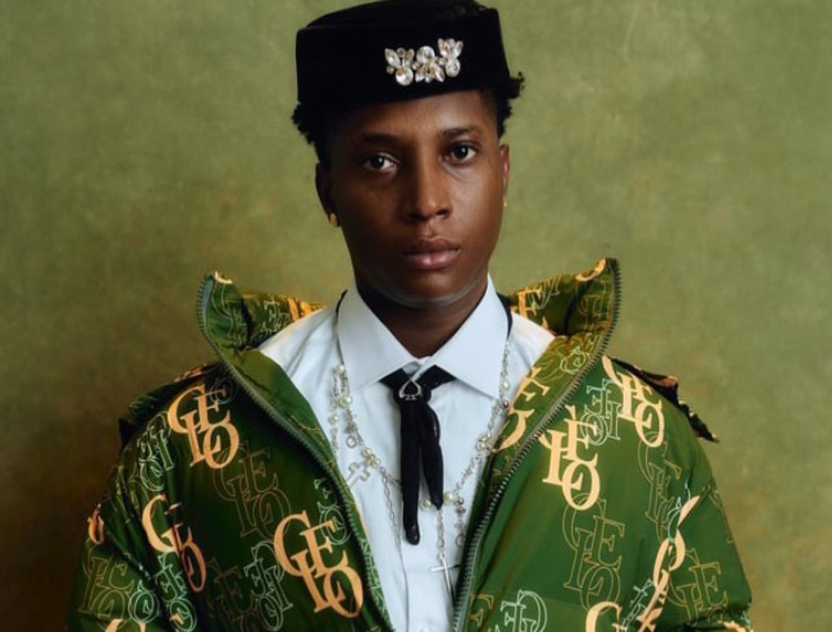 Portrait of a person wearing a green monogram jacket, white shirt, black tie, and cross necklace, with a jeweled hat motif against a green backdrop.