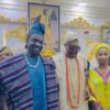 Three people in traditional Nigerian attire posing together at a ceremonial event in a richly decorated room with framed portraits behind them.