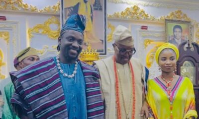 Three people in traditional Nigerian attire posing together at a ceremonial event in a richly decorated room with framed portraits behind them.