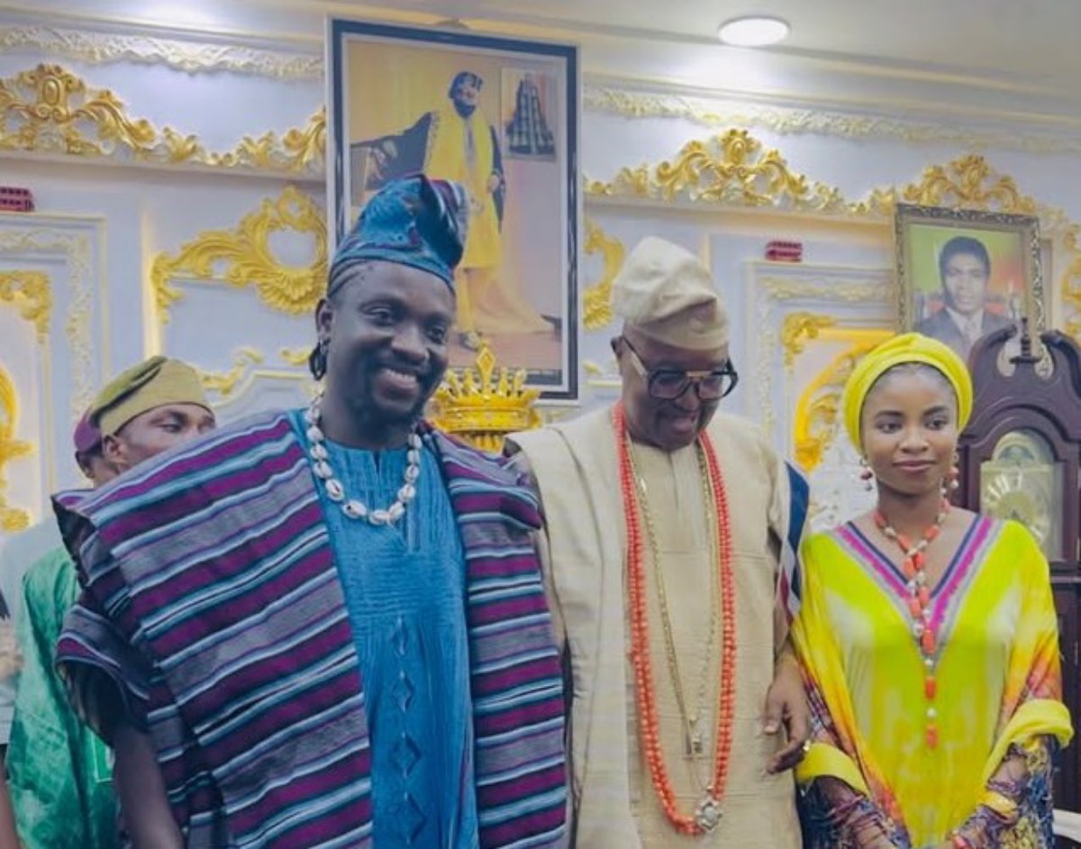Three people in traditional Nigerian attire posing together at a ceremonial event in a richly decorated room with framed portraits behind them.