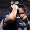 Two soccer players embrace in a celebratory hug on the pitch; the back of one jersey reads MESSI.