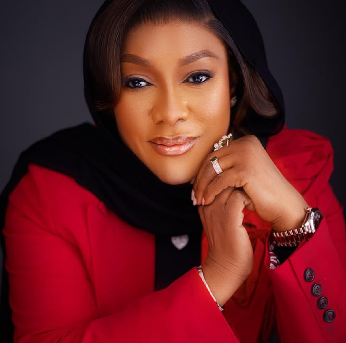 Close-up portrait of a woman in a red blazer and black hijab, resting her chin on clasped hands, wearing a watch and rings.