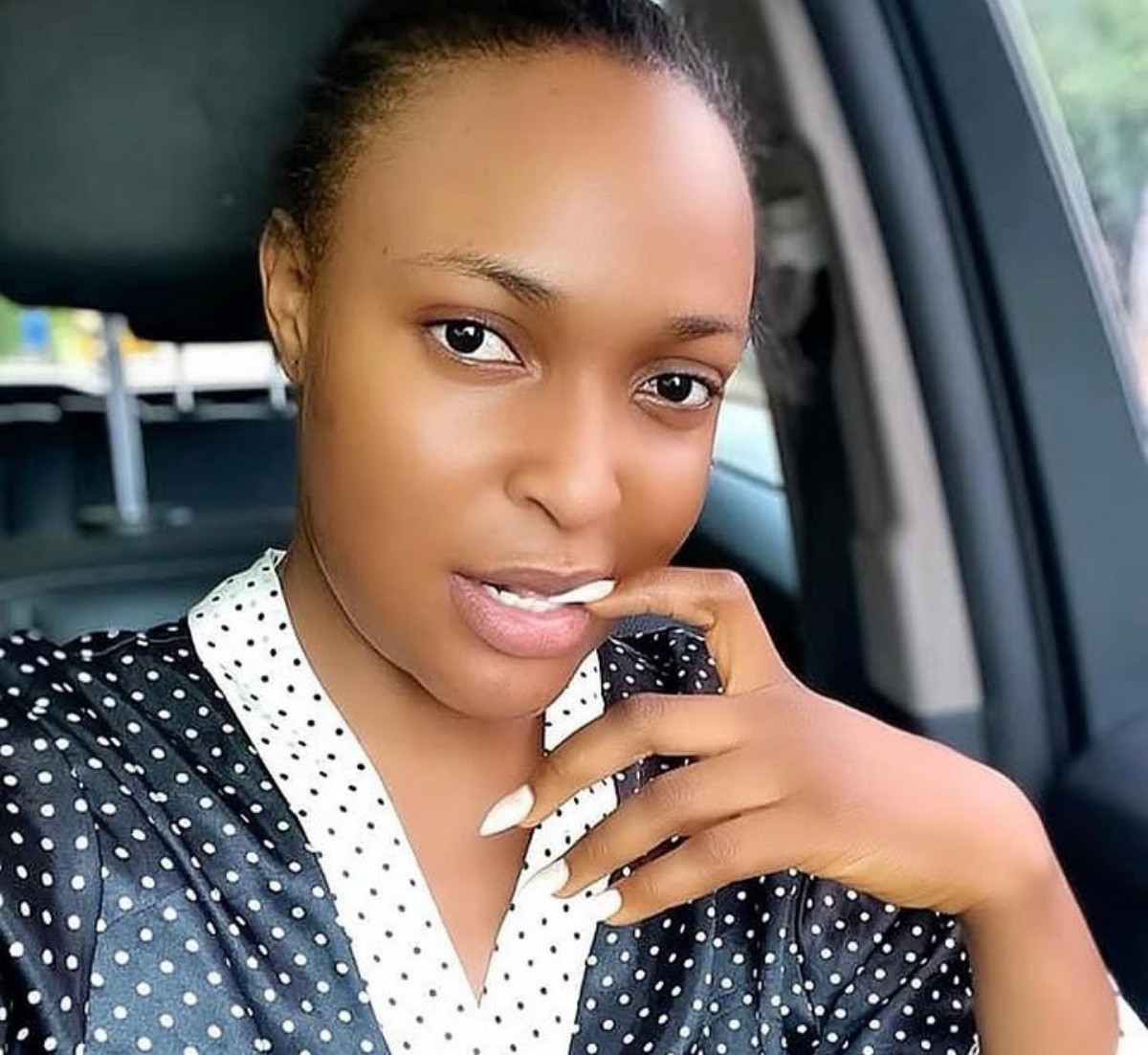 Close-up of a woman sitting in a car, wearing a black and white polka dot blouse, with her hand near her mouth and a soft smile toward the camera.
