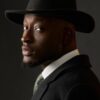 Portrait of a man wearing a black fedora and dark suit, turned to the side and looking toward the camera against a dark background.