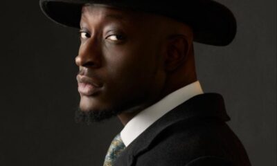 Portrait of a man wearing a black fedora and dark suit, turned to the side and looking toward the camera against a dark background.