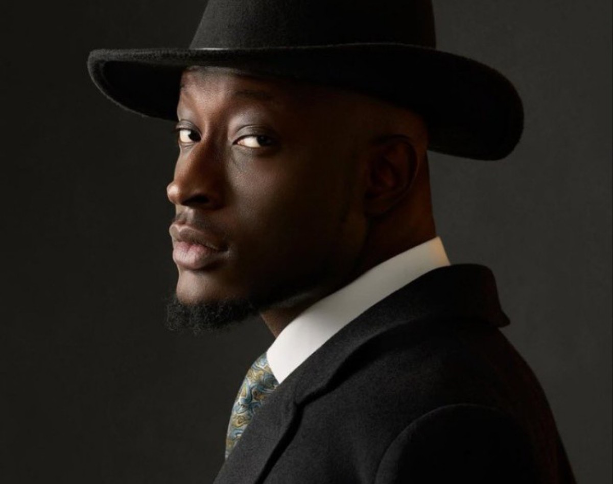 Portrait of a man wearing a black fedora and dark suit, turned to the side and looking toward the camera against a dark background.