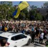 Large crowd of protesters in a city square, many holding signs; a blue and yellow Ukrainian flag waves overhead.
