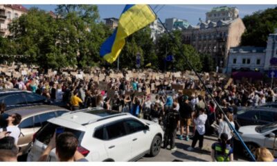 Large crowd of protesters in a city square, many holding signs; a blue and yellow Ukrainian flag waves overhead.