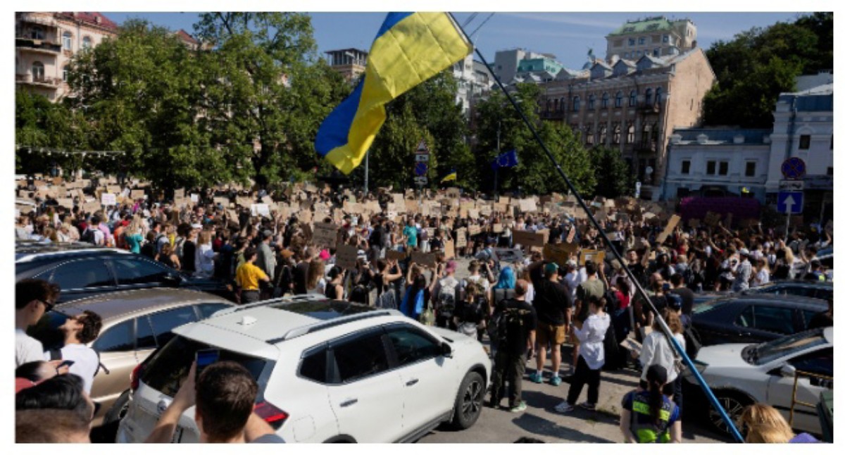 Large crowd of protesters in a city square, many holding signs; a blue and yellow Ukrainian flag waves overhead.