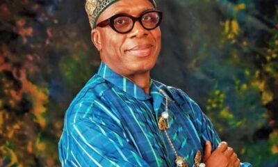 Man in ornate gold crown and blue patterned traditional attire poses with arms crossed, smiling at the camera against a colorful backdrop.