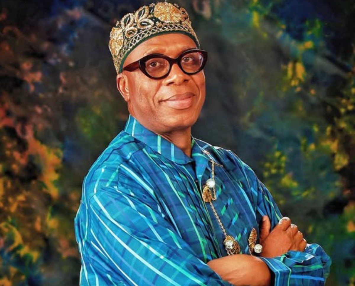 Man in ornate gold crown and blue patterned traditional attire poses with arms crossed, smiling at the camera against a colorful backdrop.