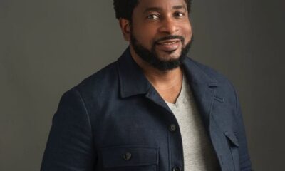 Smiling man with a beard wearing a navy jacket and gray shirt against a neutral background, posing for a portrait, head tilted slightly.
