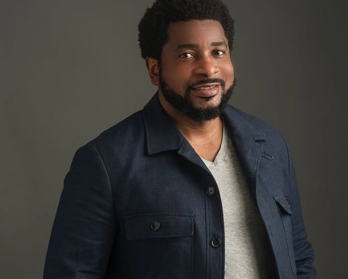 Smiling man with a beard wearing a navy jacket and gray shirt against a neutral background, posing for a portrait, head tilted slightly.