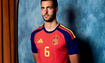 Male model wearing a red Spain national team jersey (number 6) with blue sleeves, standing against a blue textured backdrop.