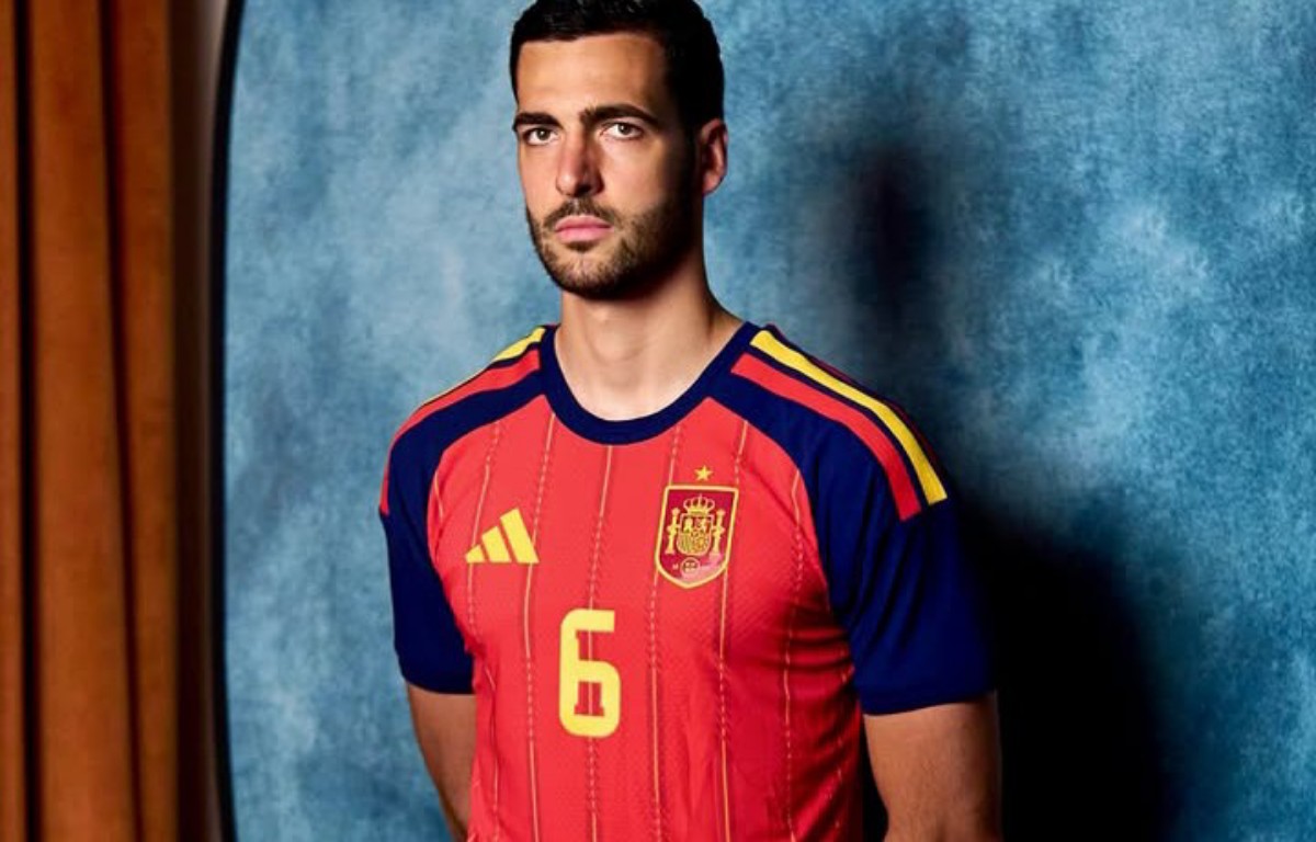 Male model wearing a red Spain national team jersey (number 6) with blue sleeves, standing against a blue textured backdrop.