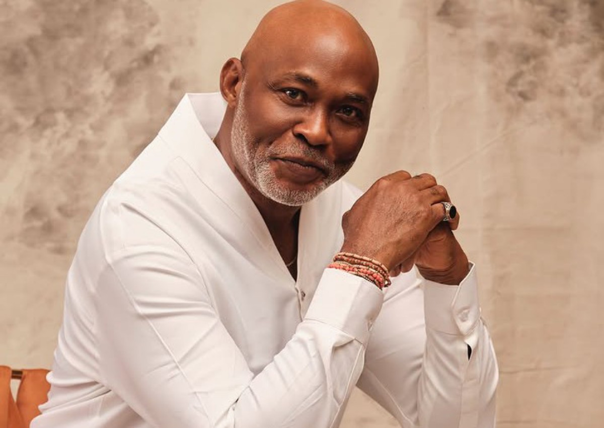 Portrait of a bald, bearded man in a white shirt, clasping his hands and looking at the camera with a gentle smile.