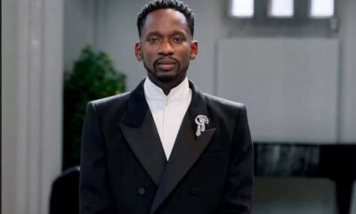 Man in a black tuxedo with a white shirt and a decorative brooch on the lapel, standing indoors with a calm expression.