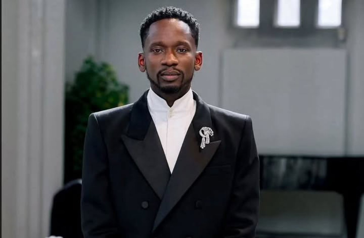 Man in a black tuxedo with a white shirt and a decorative brooch on the lapel, standing indoors with a calm expression.