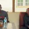 Two men smile while chatting on a couch; on the table are a soccer-ball mug, a pitcher of tea, and a bottle of water.