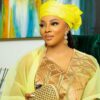 Woman in a bright yellow headwrap and sheer shawl, wearing a gold-patterned dress, holding a woven clutch on a green velvet sofa.