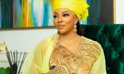 Woman in a bright yellow headwrap and sheer shawl, wearing a gold-patterned dress, holding a woven clutch on a green velvet sofa.