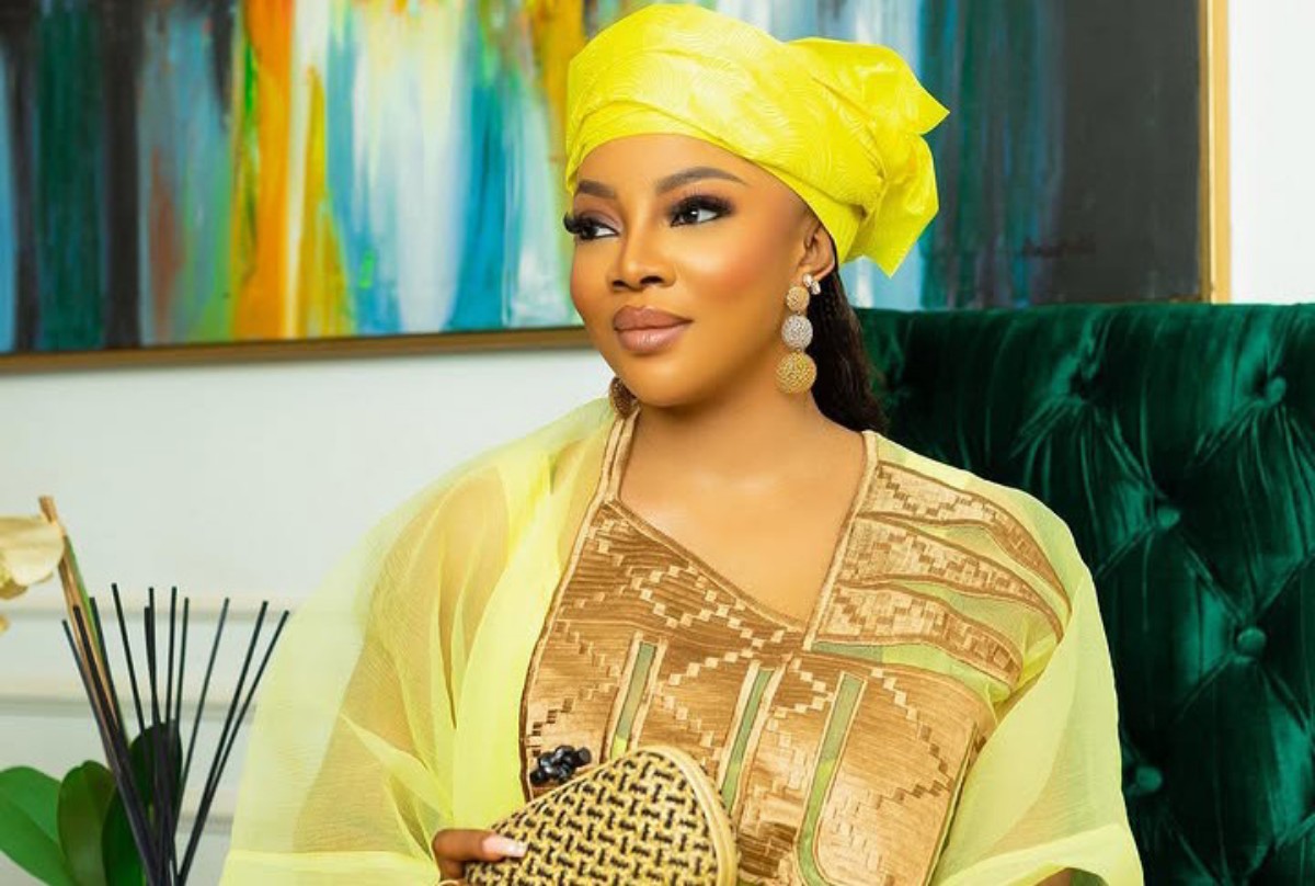 Woman in a bright yellow headwrap and sheer shawl, wearing a gold-patterned dress, holding a woven clutch on a green velvet sofa.