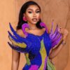 Glamorous woman wearing a multicolor beaded gown with dramatic blue and purple feather-like shoulders, pink earrings, and bold makeup.
