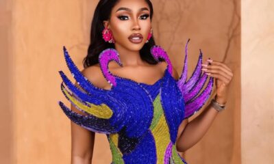 Glamorous woman wearing a multicolor beaded gown with dramatic blue and purple feather-like shoulders, pink earrings, and bold makeup.
