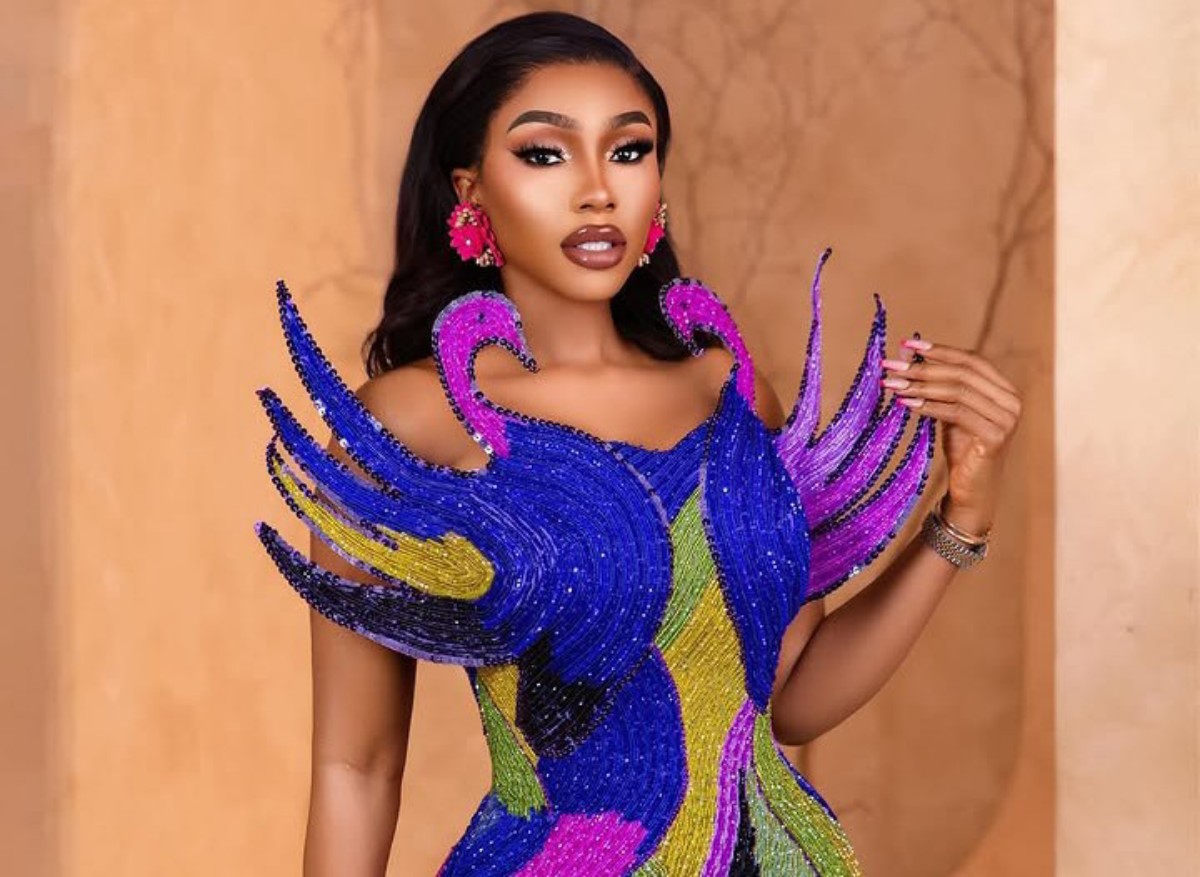 Glamorous woman wearing a multicolor beaded gown with dramatic blue and purple feather-like shoulders, pink earrings, and bold makeup.