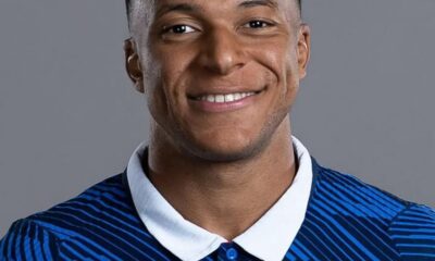 Smiling man wearing a blue striped football jersey with a white collar, featuring the France national team rooster crest and stars, against a gray background.