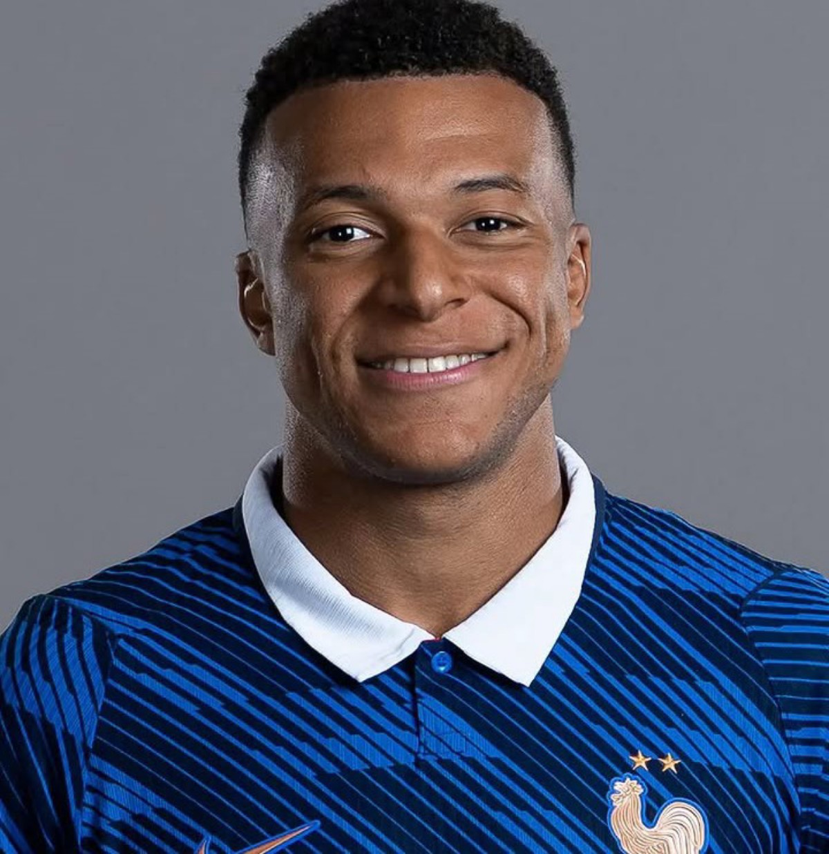 Smiling man wearing a blue striped football jersey with a white collar, featuring the France national team rooster crest and stars, against a gray background.