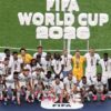 Soccer team posing on a podium with medals after winning the FIFA World Cup 2026 on a grass field.