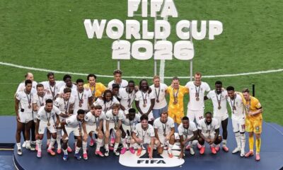 Soccer team posing on a podium with medals after winning the FIFA World Cup 2026 on a grass field.