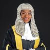 Female judge in a black robe with gold trim and a white lace jabot, wearing a white powdered wig and glasses, smiling against a dark background