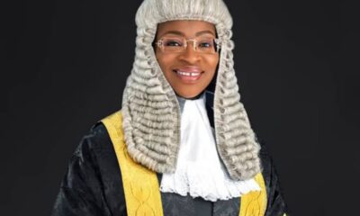 Female judge in a black robe with gold trim and a white lace jabot, wearing a white powdered wig and glasses, smiling against a dark background