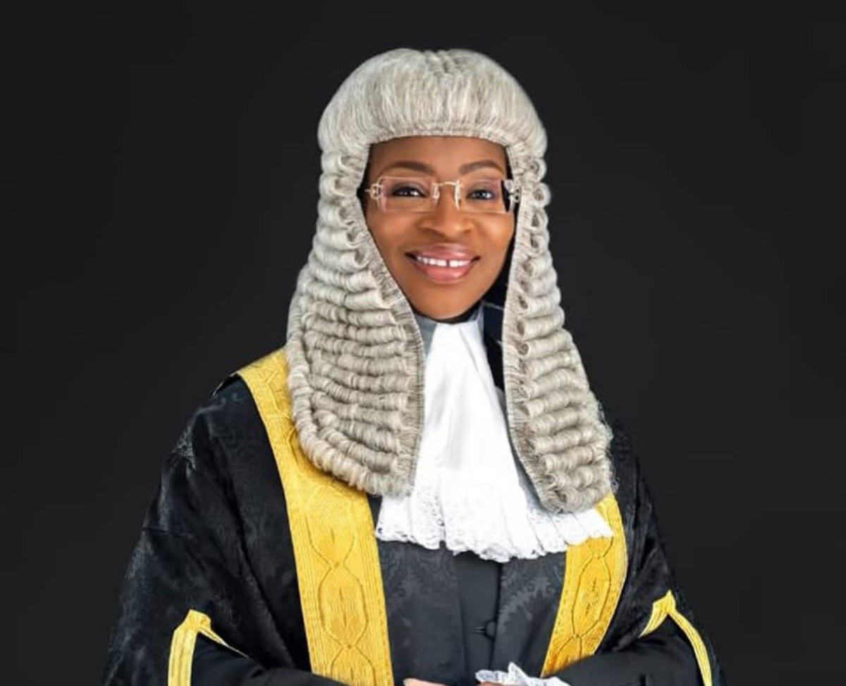 Female judge in a black robe with gold trim and a white lace jabot, wearing a white powdered wig and glasses, smiling against a dark background