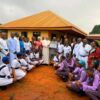 Community leaders, priests, students in uniforms pose outside a yellow building as a donor presents a check during a ceremony.