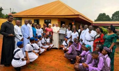 Community leaders, priests, students in uniforms pose outside a yellow building as a donor presents a check during a ceremony.