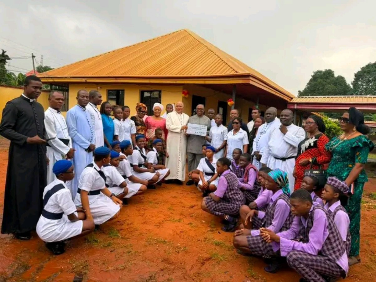 Community leaders, priests, students in uniforms pose outside a yellow building as a donor presents a check during a ceremony.