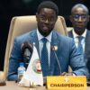 Chairperson in a blue suit speaking at a conference table with two microphones and a 'CHAIRPERSON' nameplate on the desk.