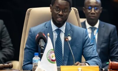 Chairperson in a blue suit speaking at a conference table with two microphones and a 'CHAIRPERSON' nameplate on the desk.