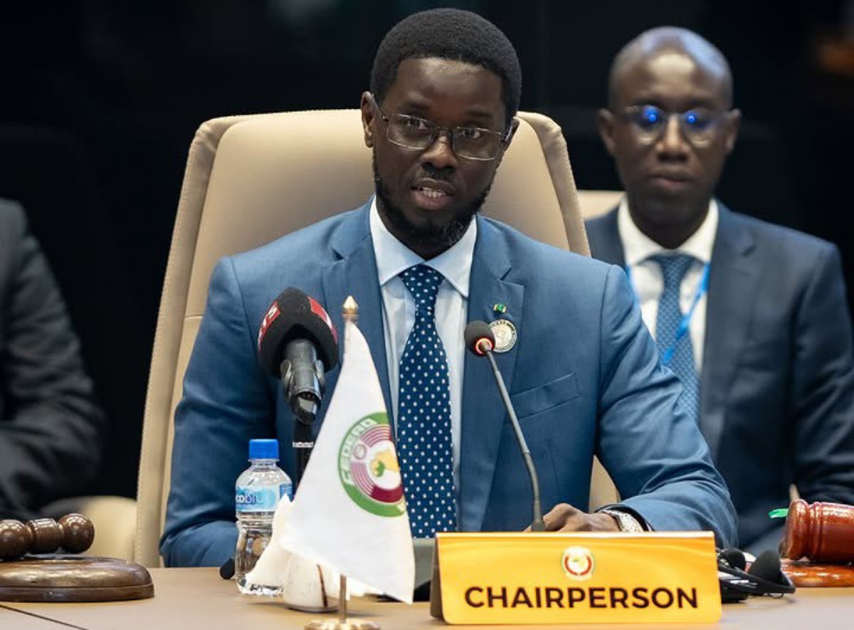 Chairperson in a blue suit speaking at a conference table with two microphones and a 'CHAIRPERSON' nameplate on the desk.