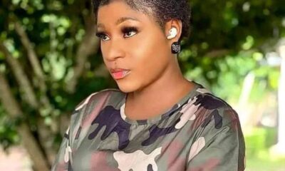 Side-profile portrait of a woman with short curly hair wearing a camouflage top, outdoors, with a wireless earbud in her left ear.