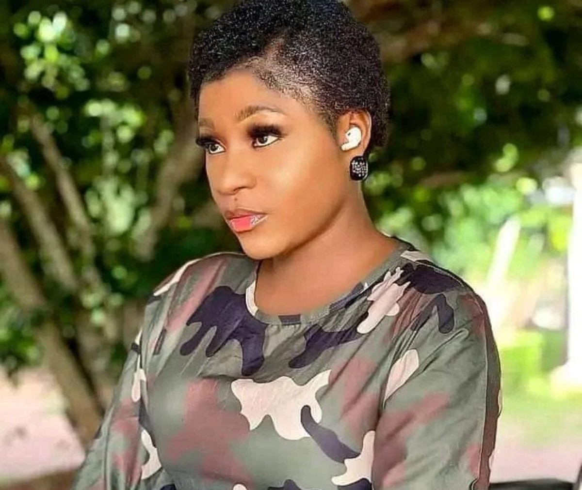 Side-profile portrait of a woman with short curly hair wearing a camouflage top, outdoors, with a wireless earbud in her left ear.