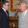 Two men in suits converse in an opulent, gold-accented room with a turquoise parrot statue on a pedestal between them.