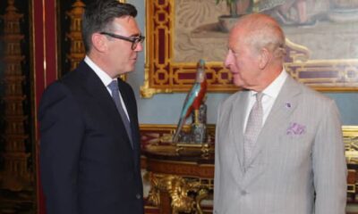 Two men in suits converse in an opulent, gold-accented room with a turquoise parrot statue on a pedestal between them.